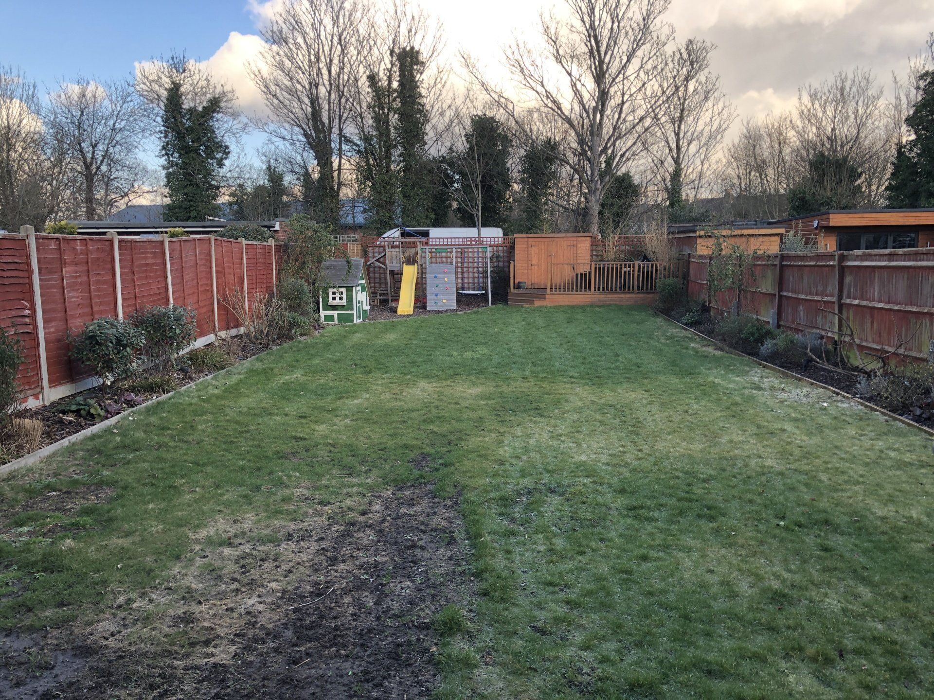 A backyard with a green lawn, wooden fence, small play structure, and shed under a cloudy sky.