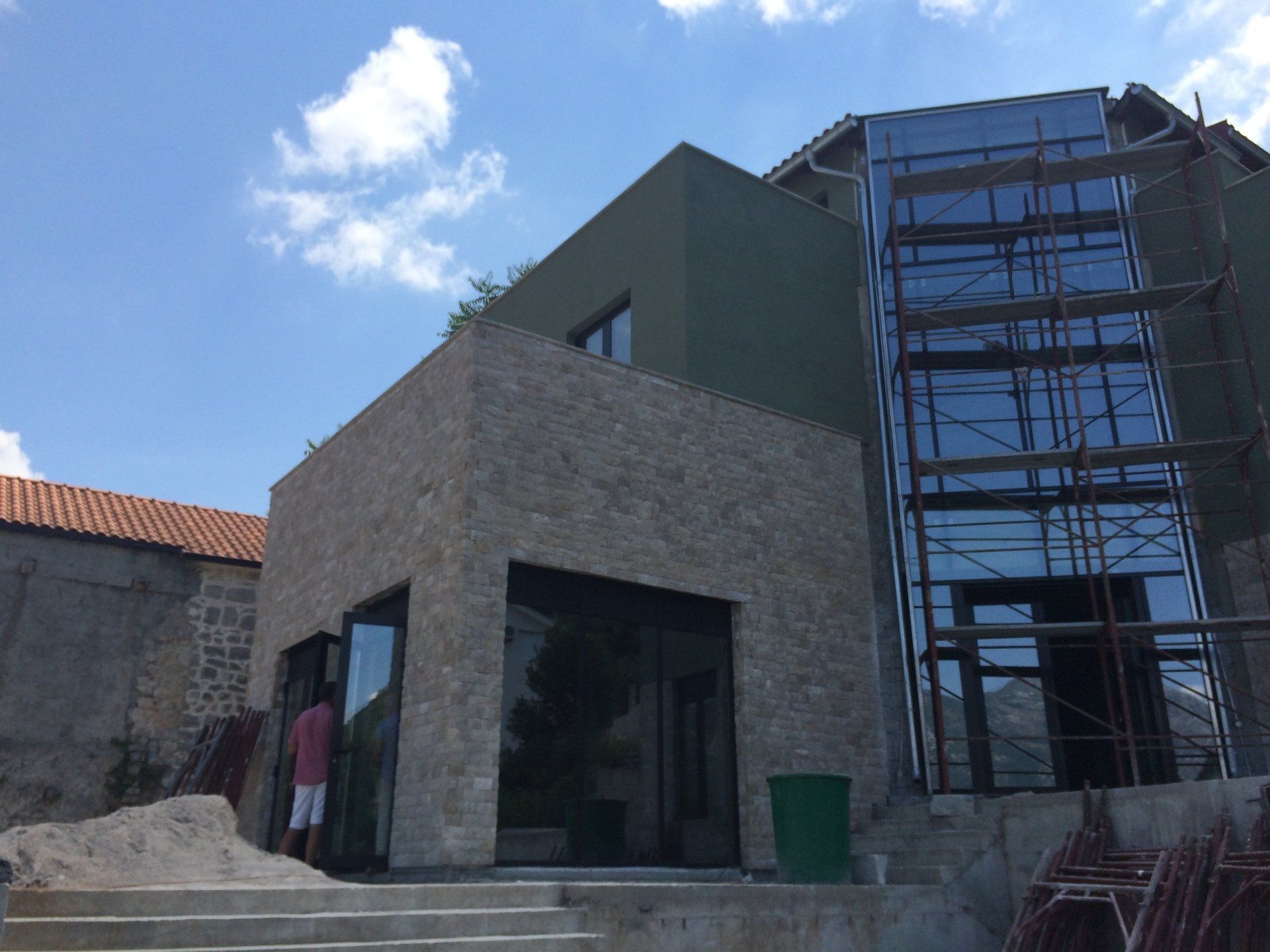 Modern building under construction with stone and glass exterior, blue sky, and a person standing near open doors.
