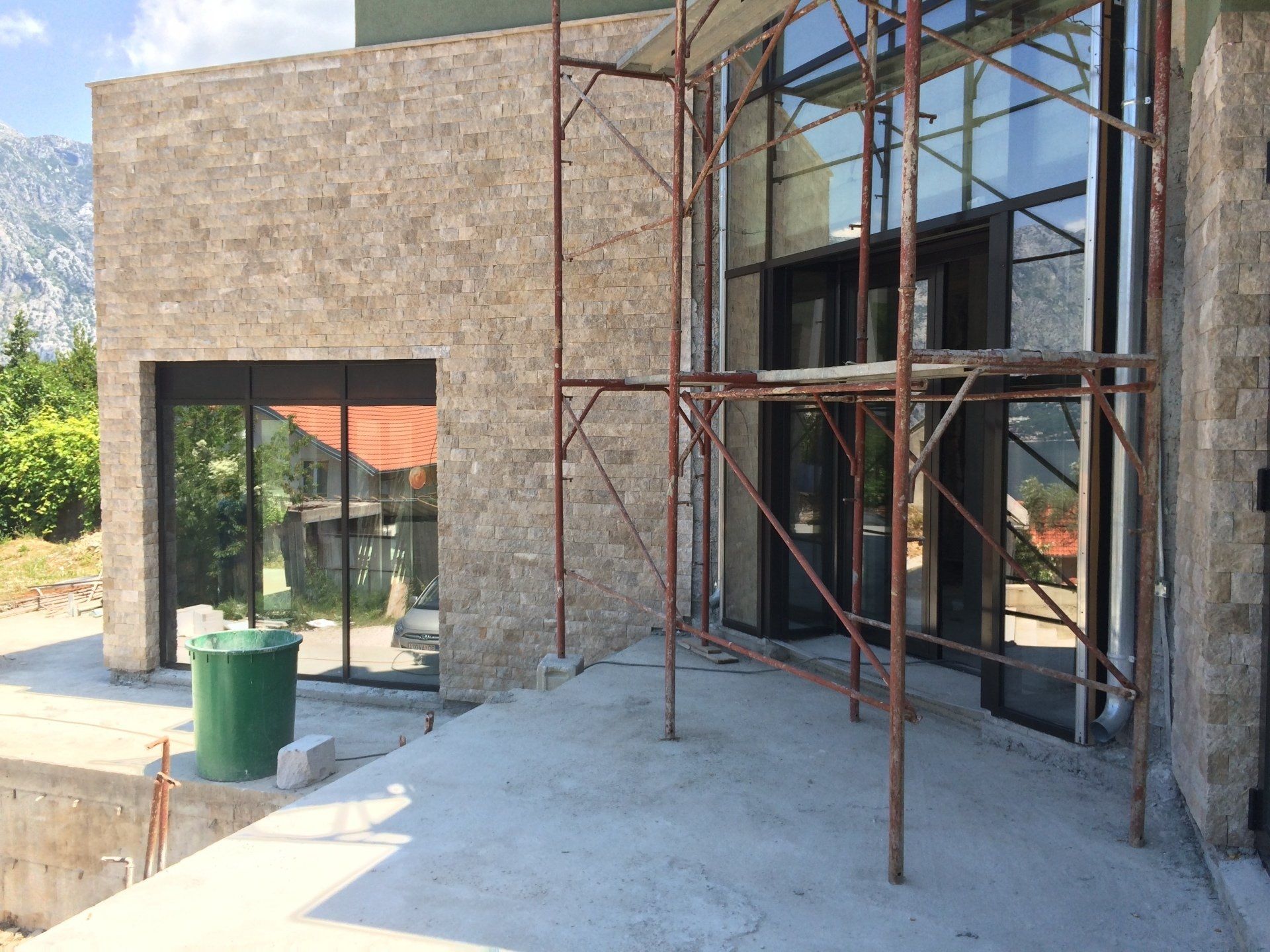 Exterior of a building with stone cladding and large glass windows; scaffolding present. A green trash can sits outside.