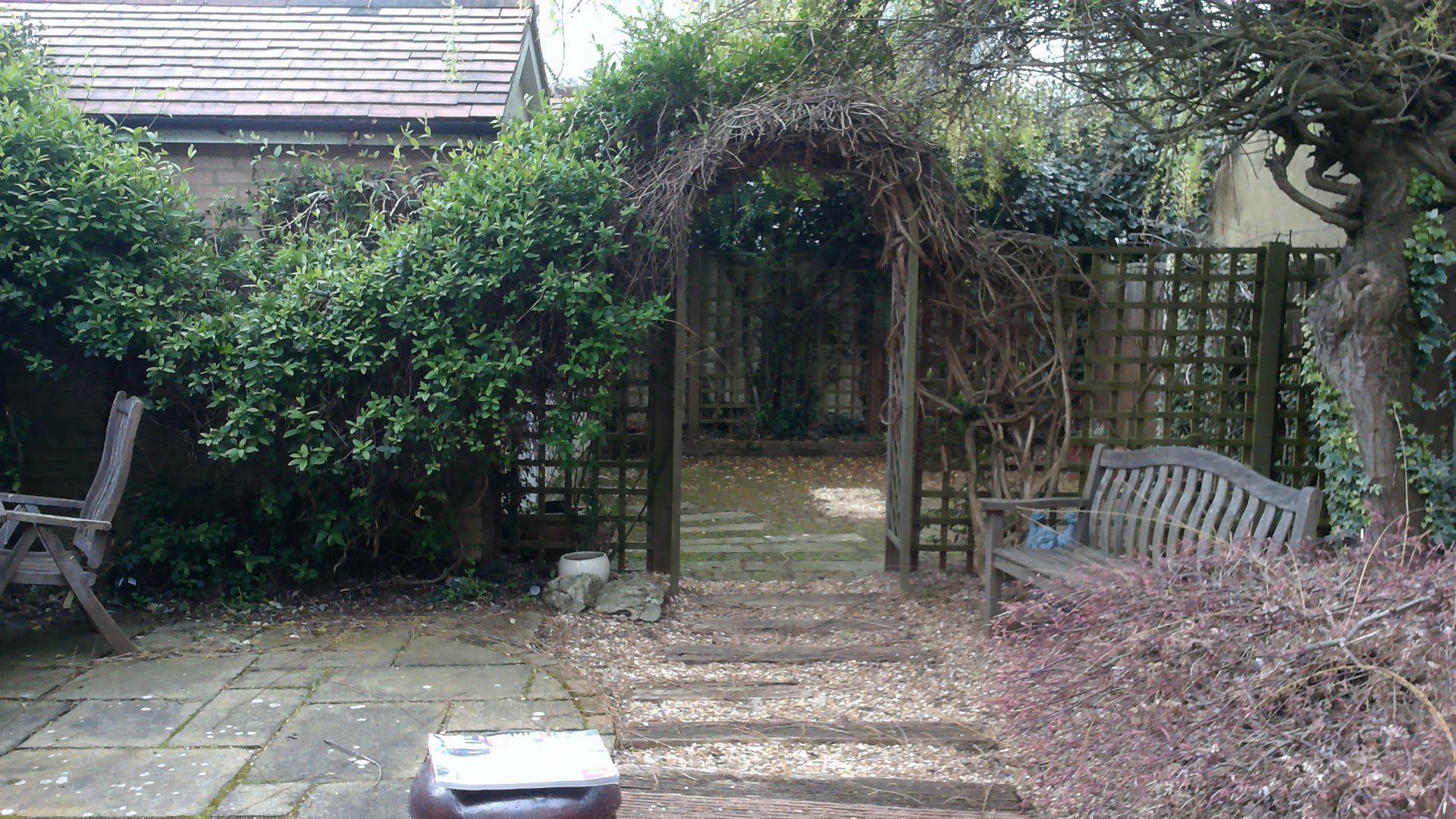 Garden path with stone steps leading through a trellis archway, flanked by benches and lush greenery.