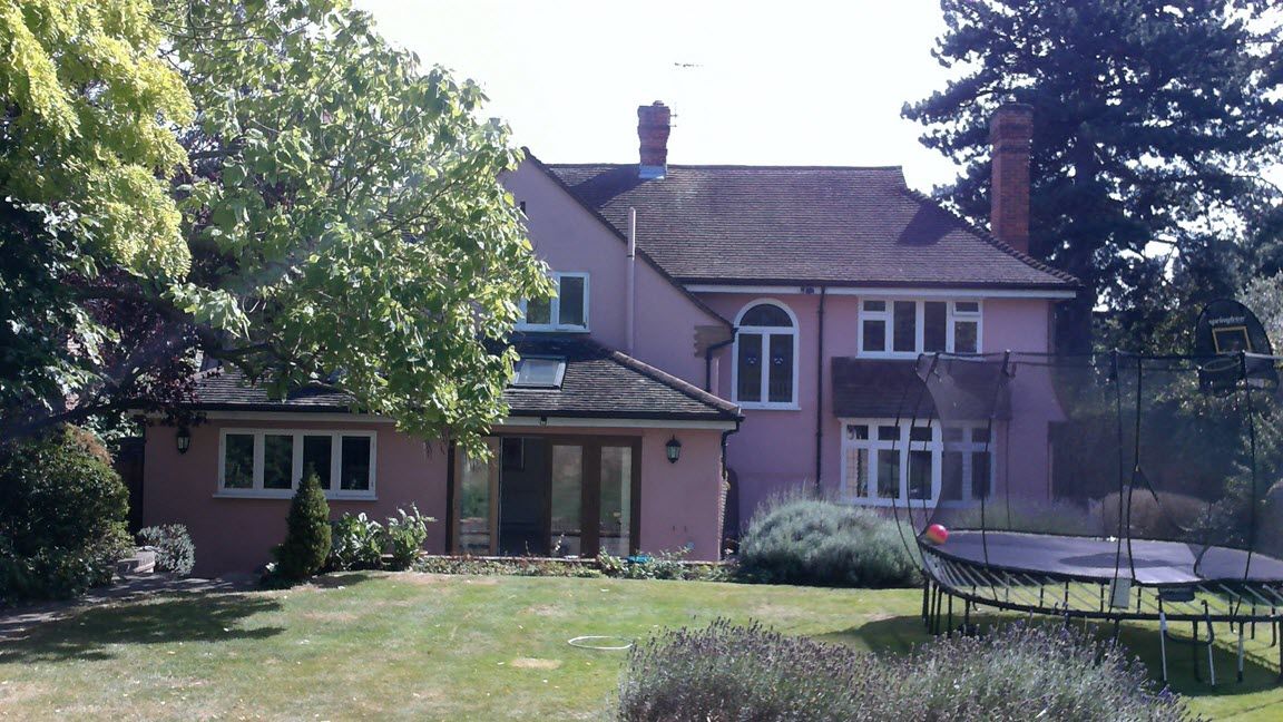 Pink house with brown roof, green lawn, trees, and a trampoline.