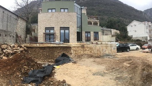 Modern house under construction with stone facade, glass windows, and dirt surrounding it.