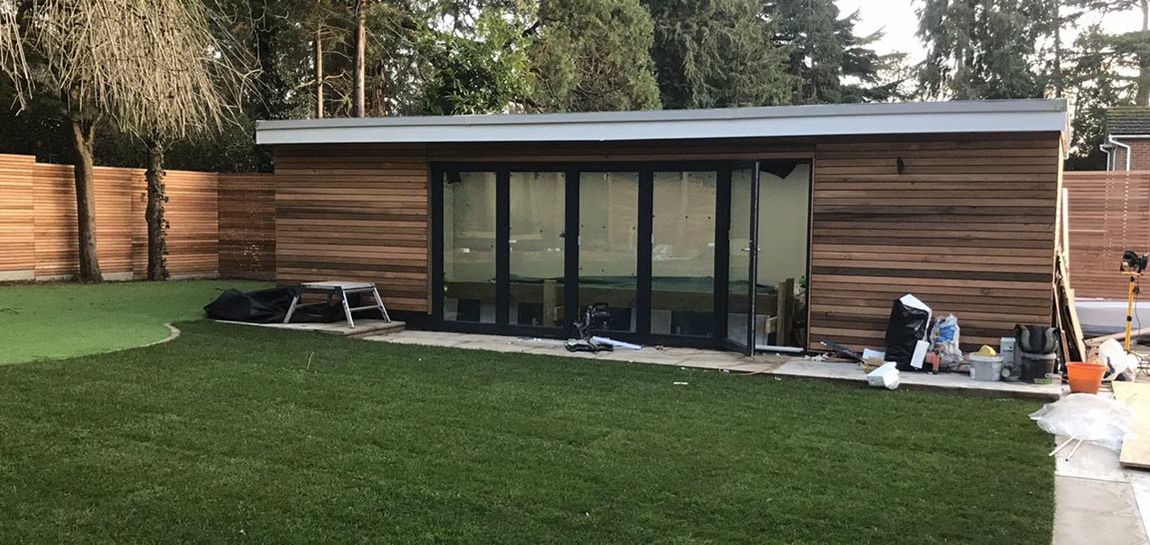 A wooden outbuilding with glass doors, set in a yard with green lawn and a wooden fence. Construction materials are visible.