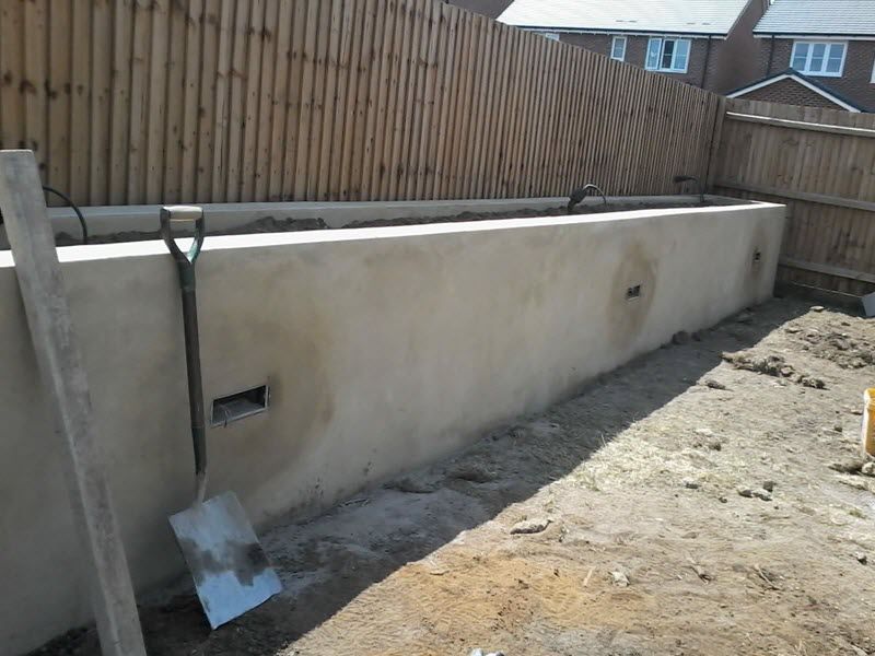 Long, plastered retaining wall under a wooden fence, with a shovel leaning against it.