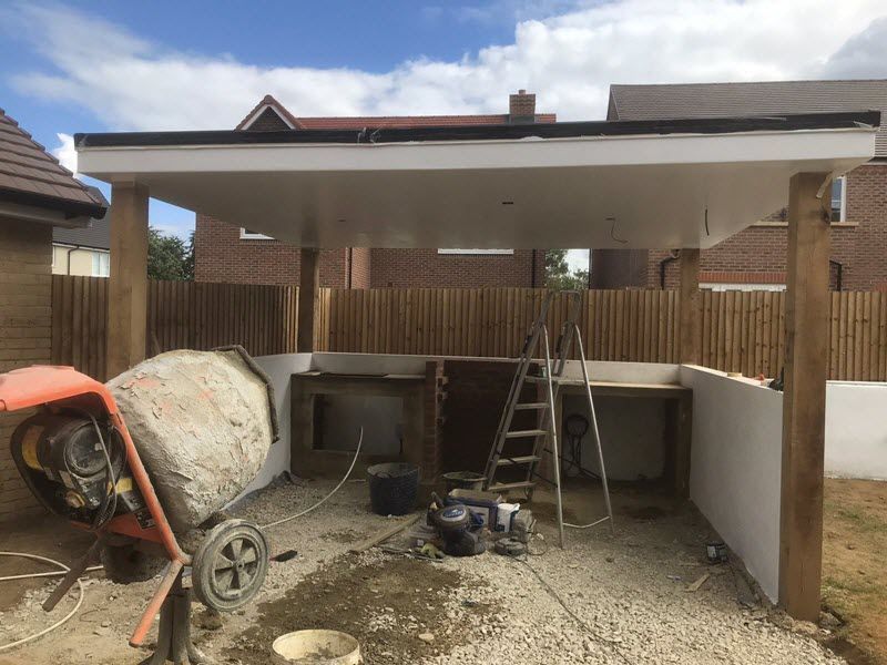 Construction of a backyard outdoor kitchen under a white roof. Cement mixer, wood supports, and tools are present.