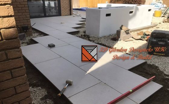 Construction of a patio with large gray tiles being laid. A level and hammer are present, along with a rectangular planter.