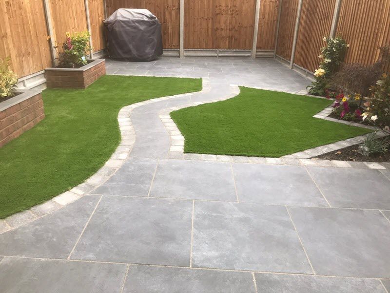 Patio with stone pavers, winding brick path, green artificial grass, and wood fence.
