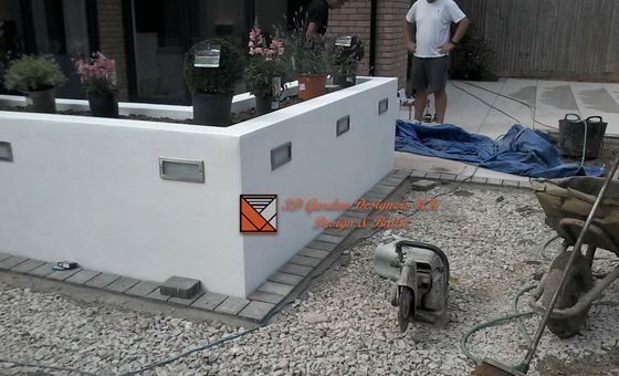 White retaining wall with inset lights, plants, gravel and patio. Construction tools are in view.