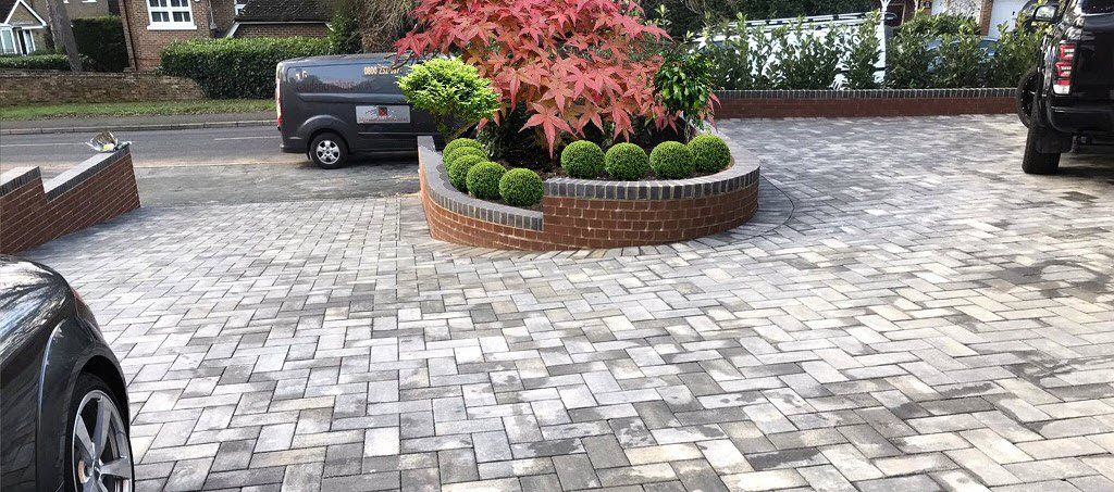 Driveway with brick pavers, central raised flower bed with plants, and parked cars.