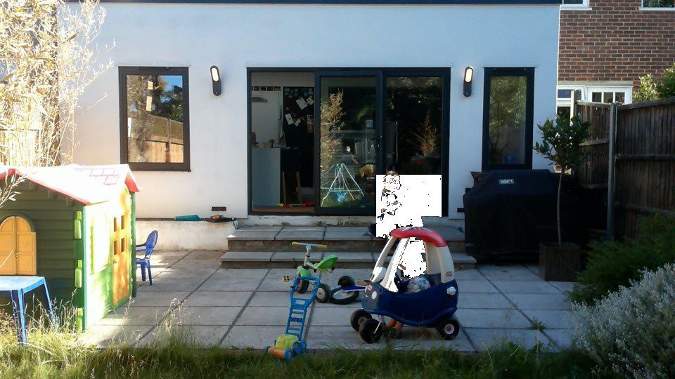 White house exterior with black-framed doors/windows, patio, and backyard toys: playhouse, slide, car.
