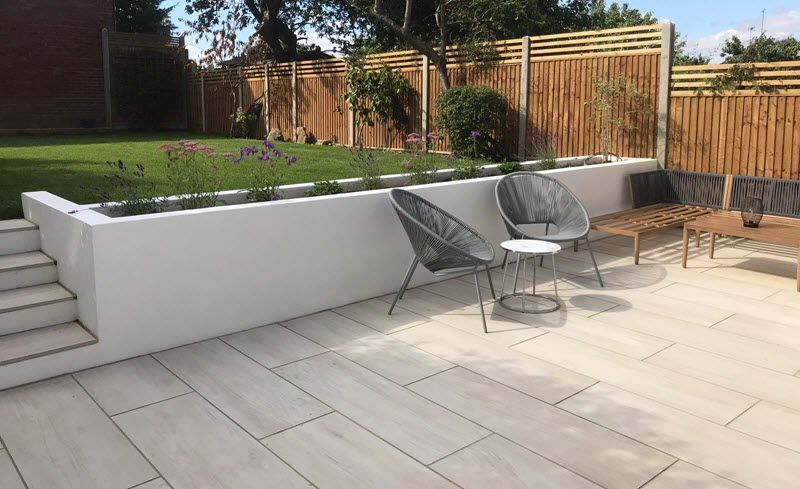 Patio with light-colored tile, a white retaining wall with flowers, and modern chairs. Bamboo fence in the background.