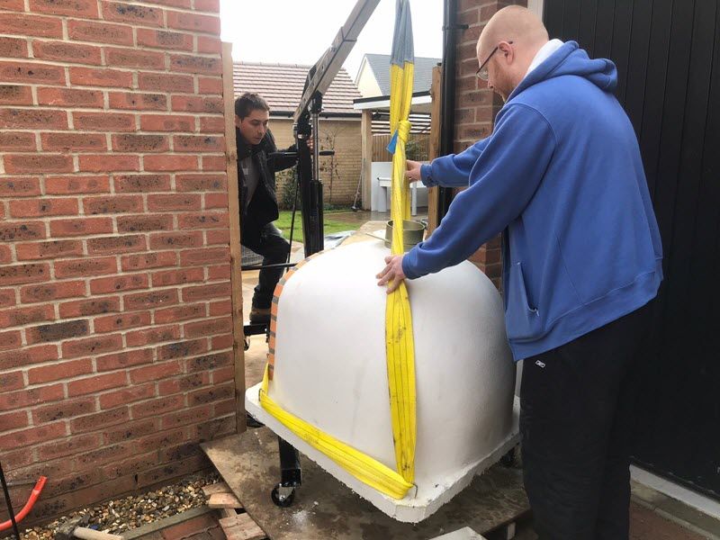 Two people using a crane to lift a large white tub near a brick wall and a garage door.