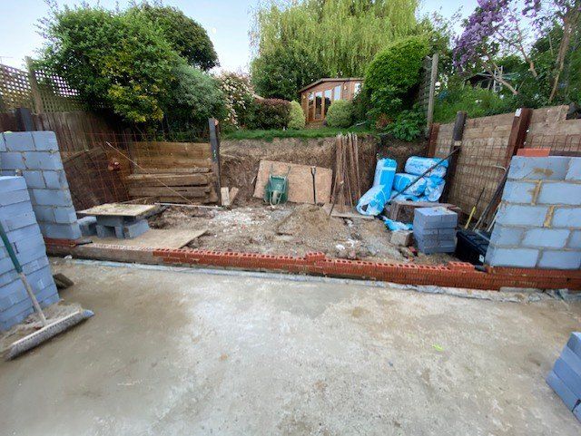 Construction site: retaining wall being built in a garden, with blocks, wood, and materials visible.