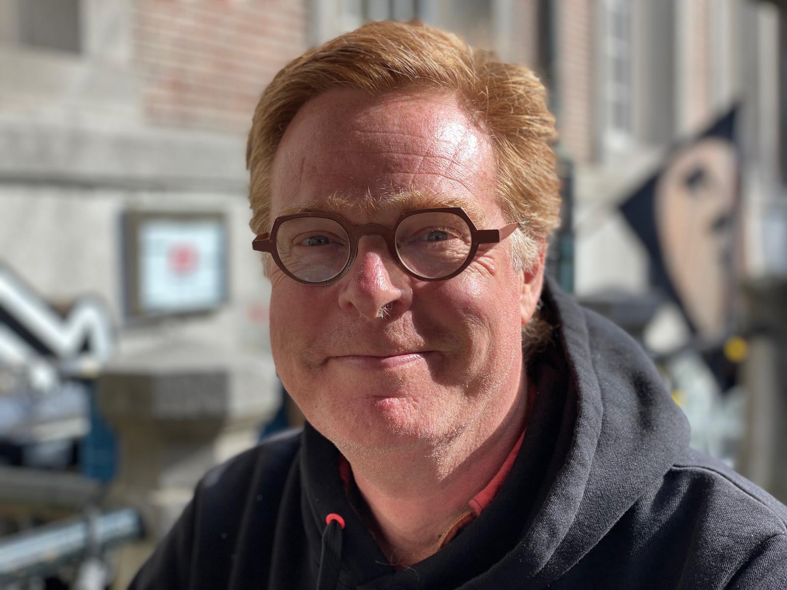 Een persoon met rood haar en een ronde bril, gekleed in een zwarte hoodie, glimlacht lichtjes tegen een wazige stedelijke achtergrond.
