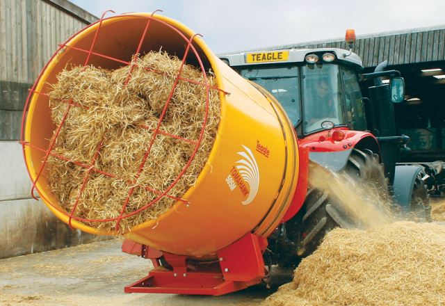 Traktor læsser halm af en gul blandevogn i en ladegård, mens halmen falder ned på jorden.