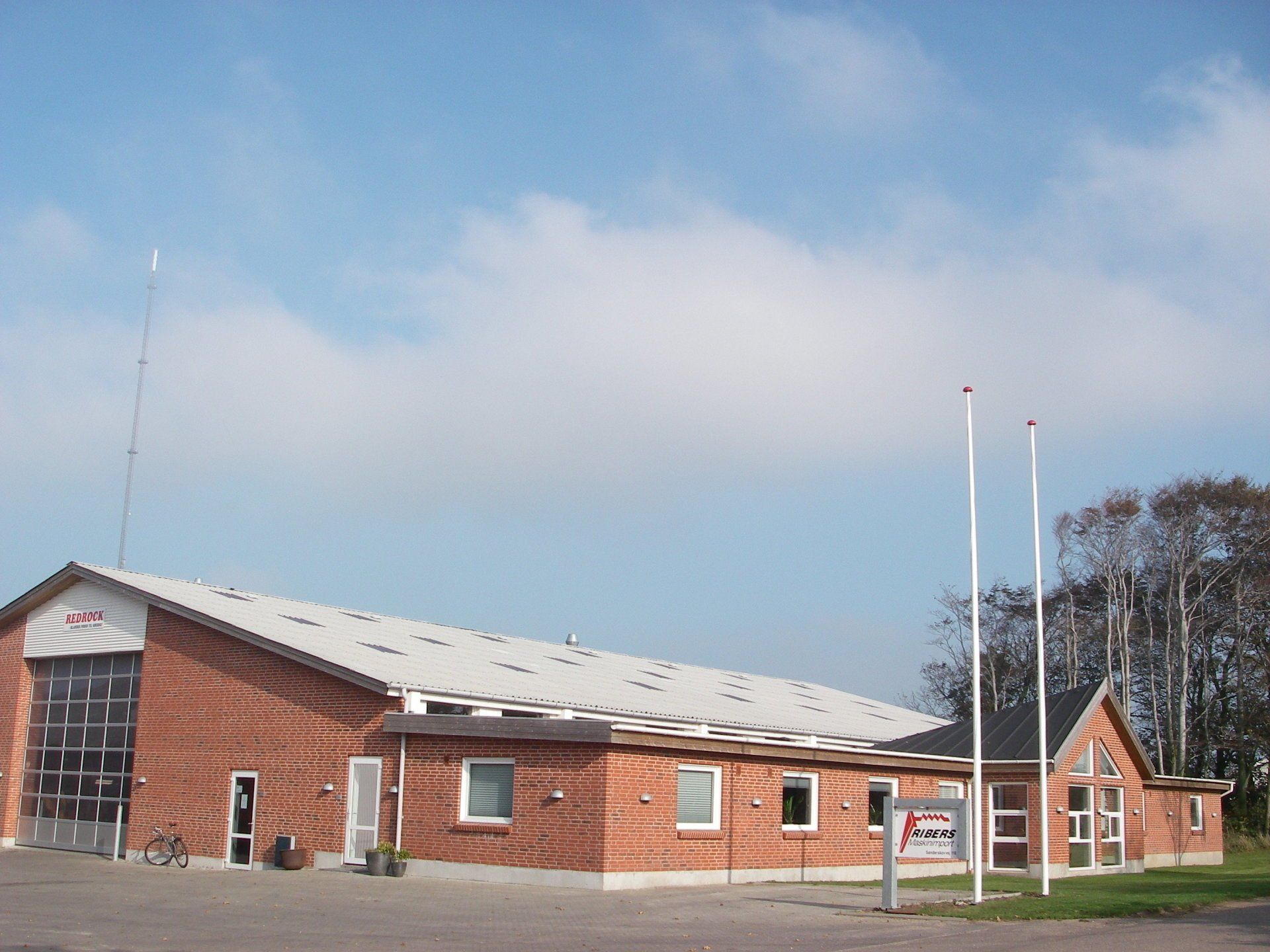 Lav murstensbygning med hvidt tag under en overskyet himmel, med flagstænger og et skilt foran