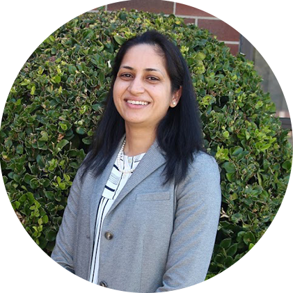 A woman in a grey jacket is smiling in front of a bush