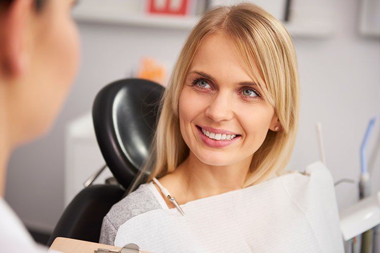 A woman is smiling while sitting in a dental chair talking to a dentist.