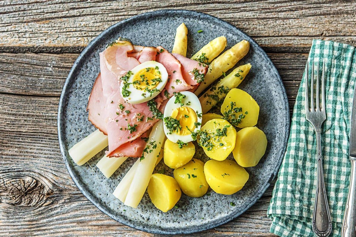 Een bord met gekookte aardappelen, ham en hardgekookte eieren op een rustieke houten tafel met een groen servet.