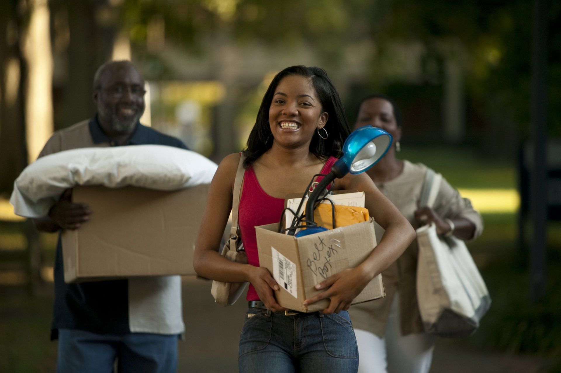 Family carrying boxes and belongings, moving into a new home; smiling young person in the center.