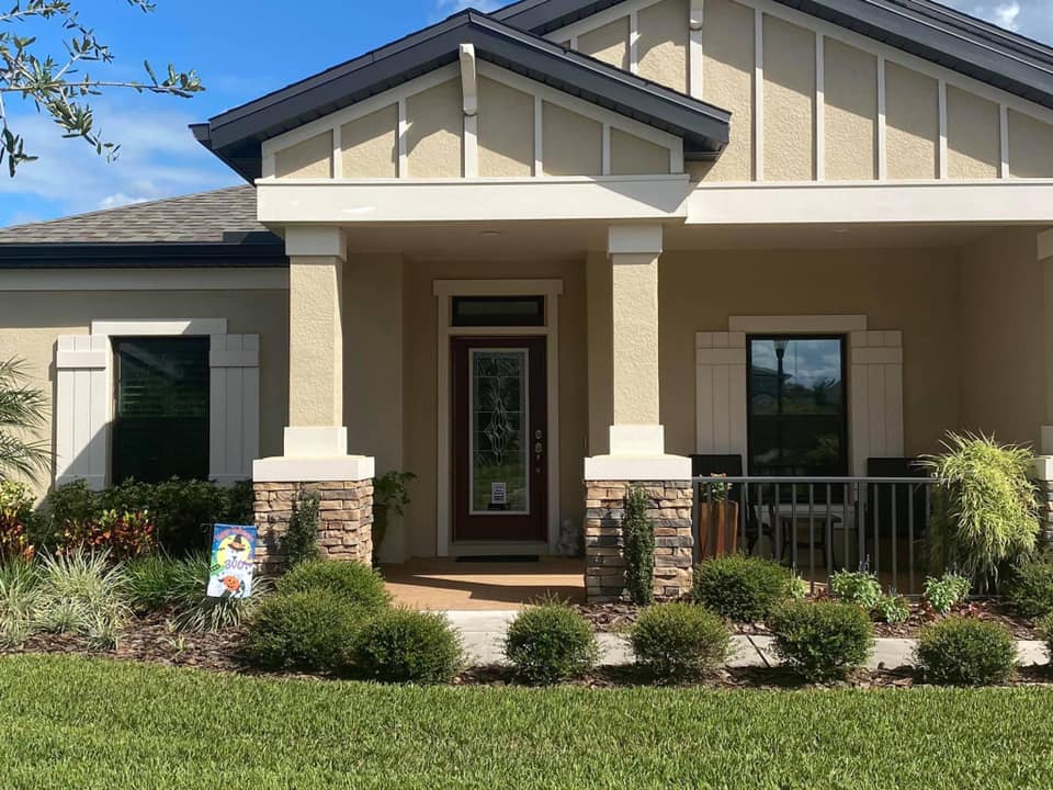 Front view of a beige house with a covered porch, white trim, stone columns, and landscaped front yard