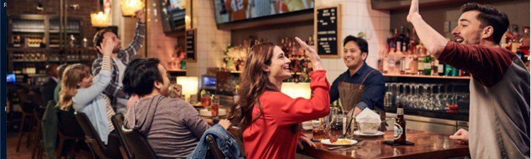A group of people are sitting at a bar raising their arms in the air.