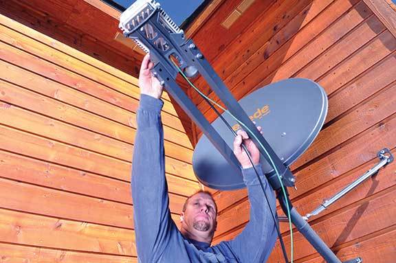 A man is installing a satellite dish on the side of a wooden building.