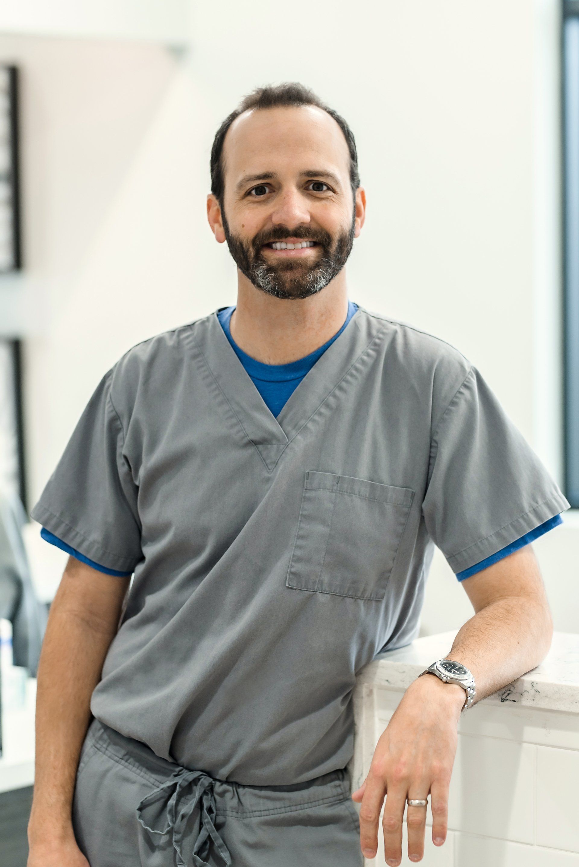 A man with a beard is wearing scrubs and standing next to a counter.