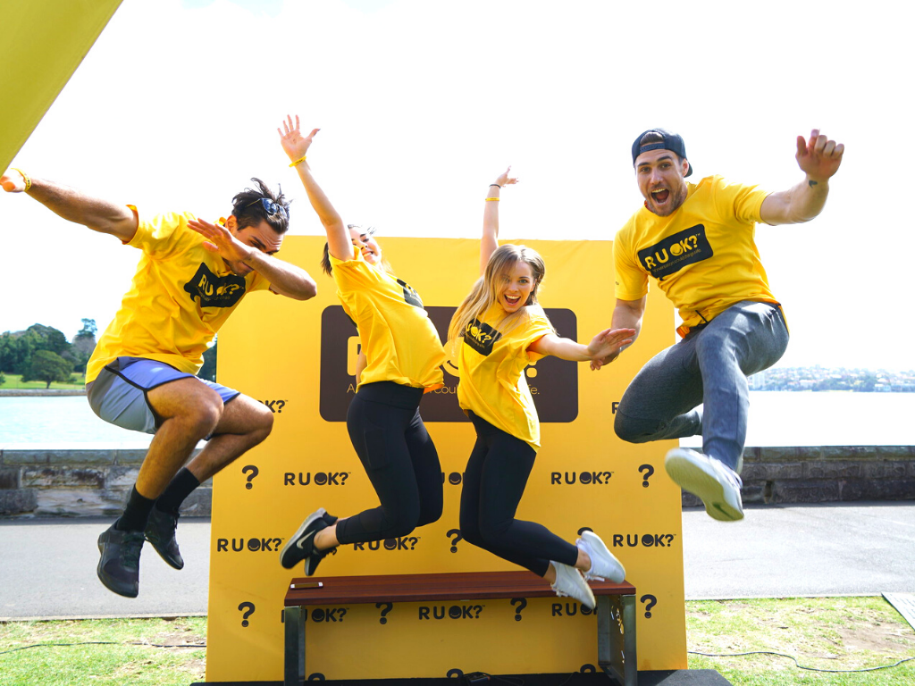 Group of excited young people jumping in front of an R U OK? Sign