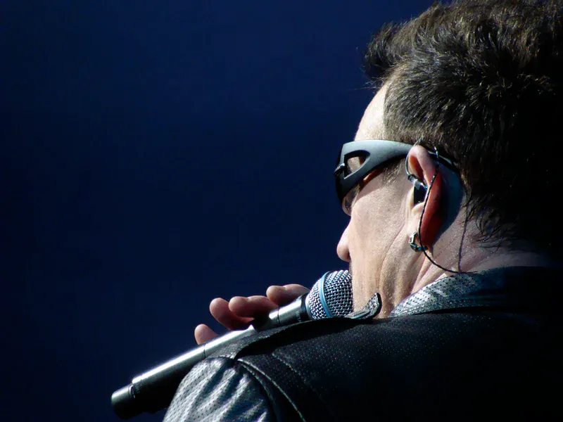 Man with microphone, wearing sunglasses and earpiece, singing on stage against a dark blue background.
