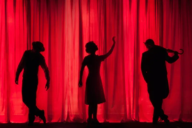 Silhouettes of three people on a stage with red curtains. One raises an arm, another holds an umbrella.