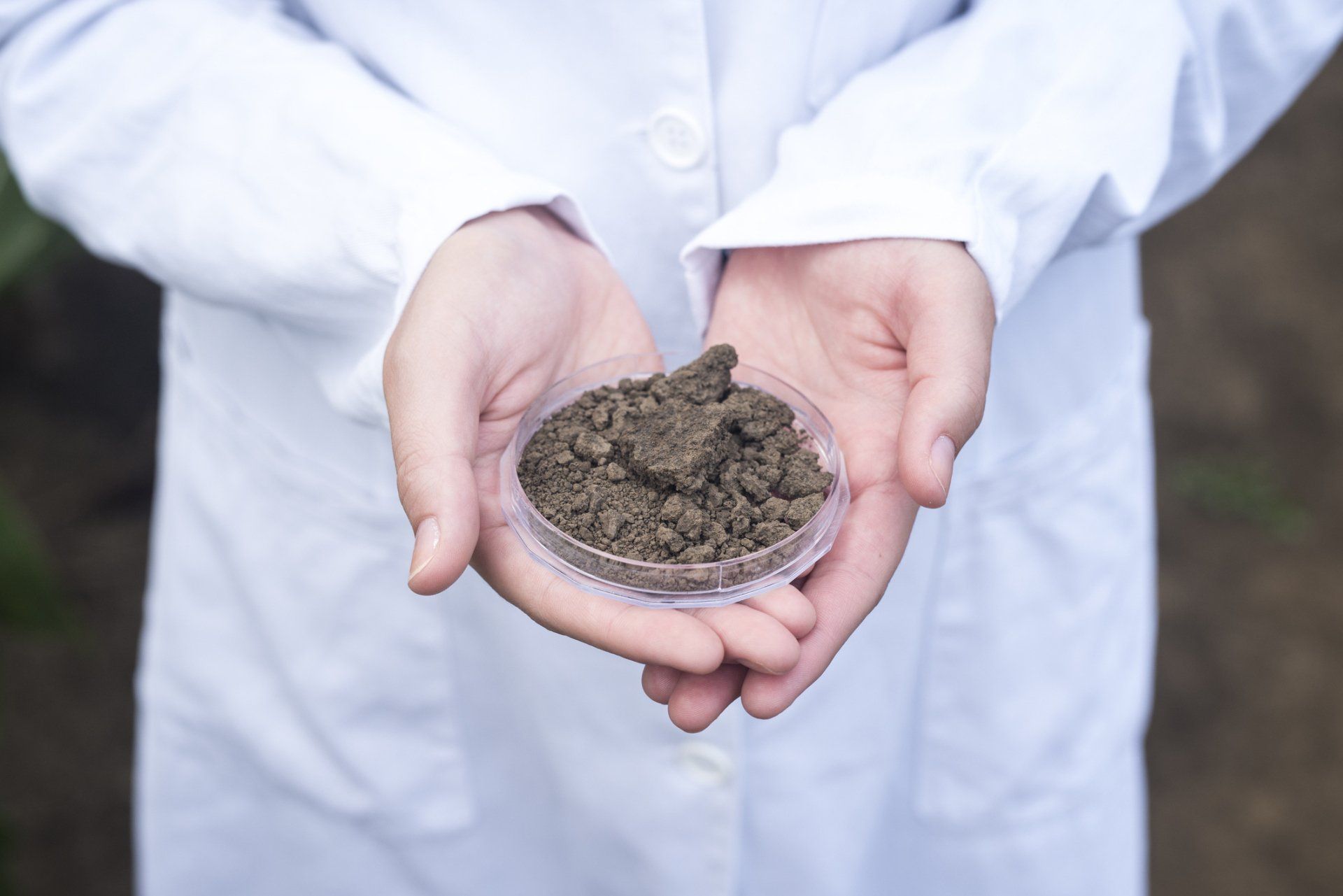 Person in lab coat holding a petri dish with soil, likely for analysis.