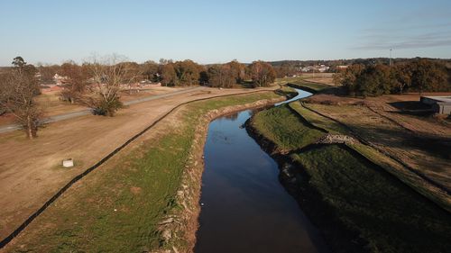 A brown river flows through grassy banks under a clear blue sky. A rural setting with trees and buildings.