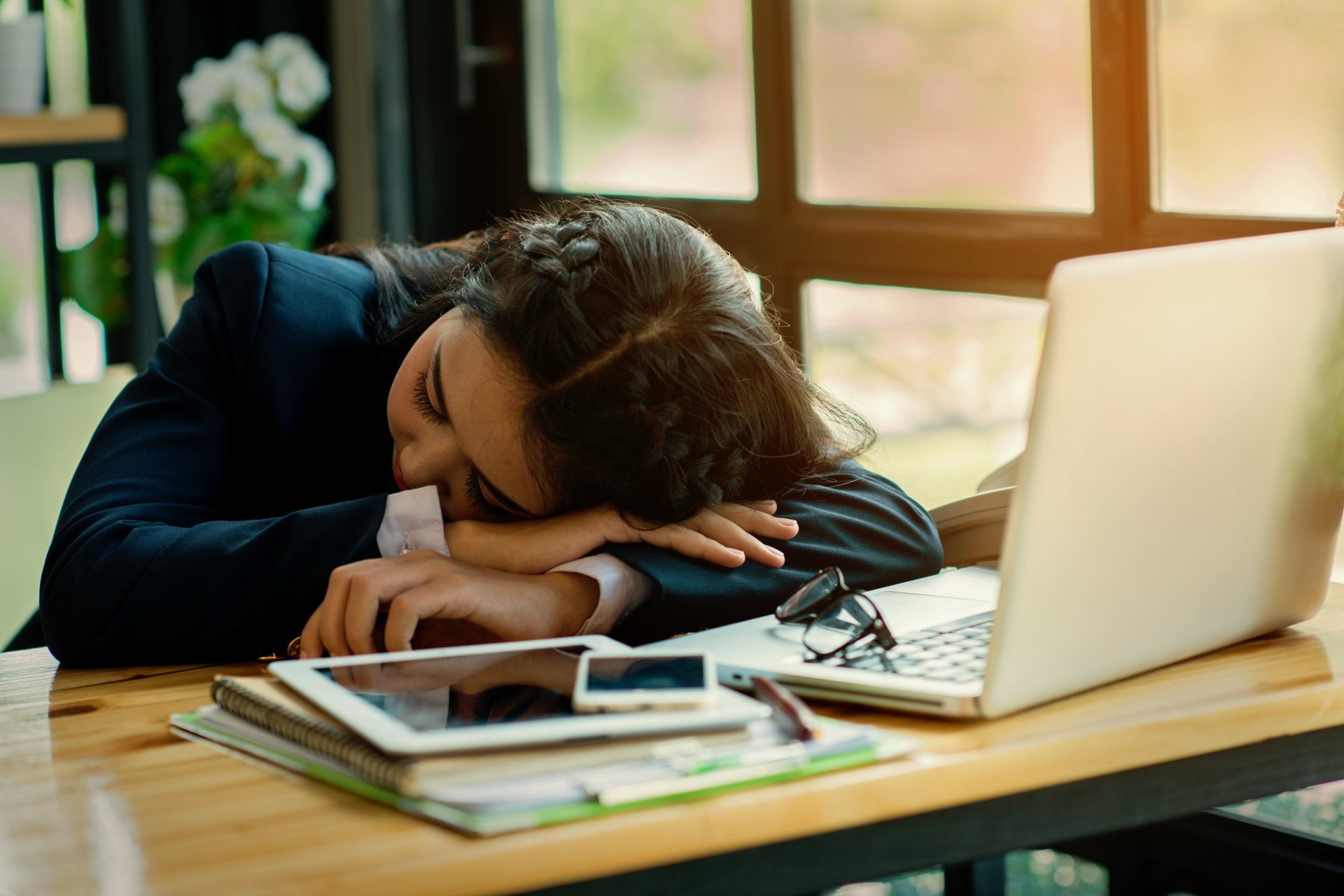 A woman is sleeping at a desk with a laptop and a tablet.