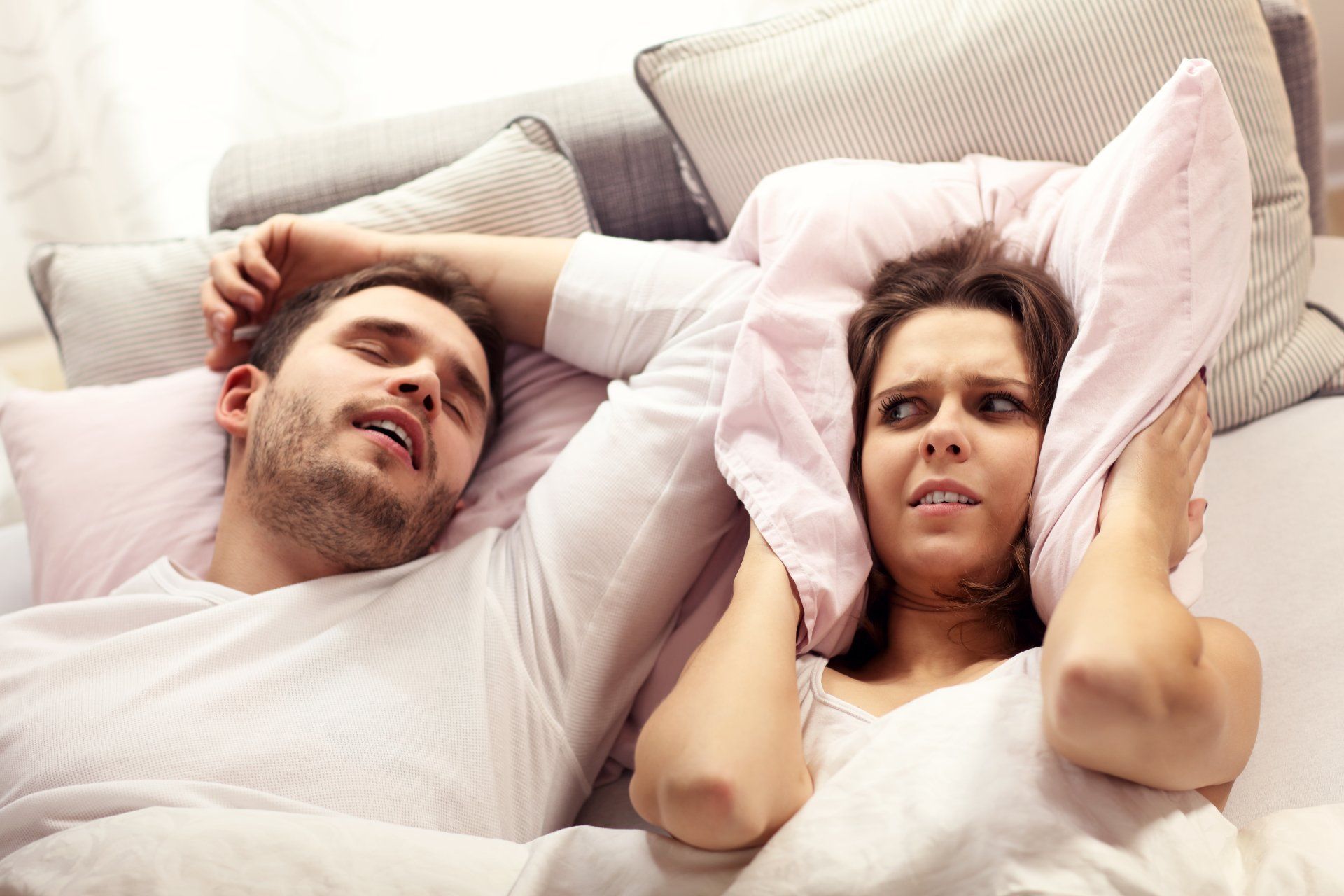 A man is snoring while a woman covers her ears with a pillow.
