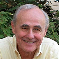 Smiling man with gray hair in a pale yellow shirt, outdoors against leafy greenery