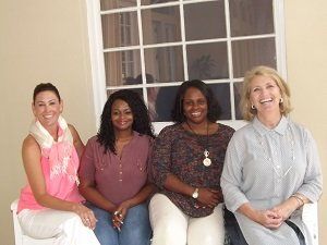 Four women are sitting on a bench in front of a window and smiling.