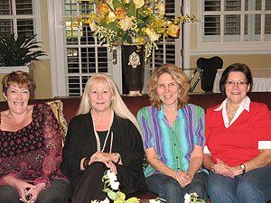 Four women are sitting on a couch in front of a vase of flowers.