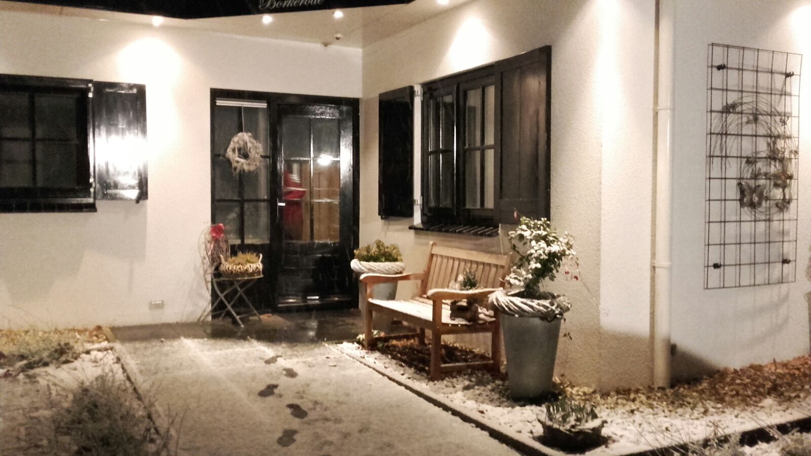 Besneeuwde buitenkant van een gebouw met een zwart omlijste deur en luiken, een bankje en planten. Voetafdrukken in de sneeuw.