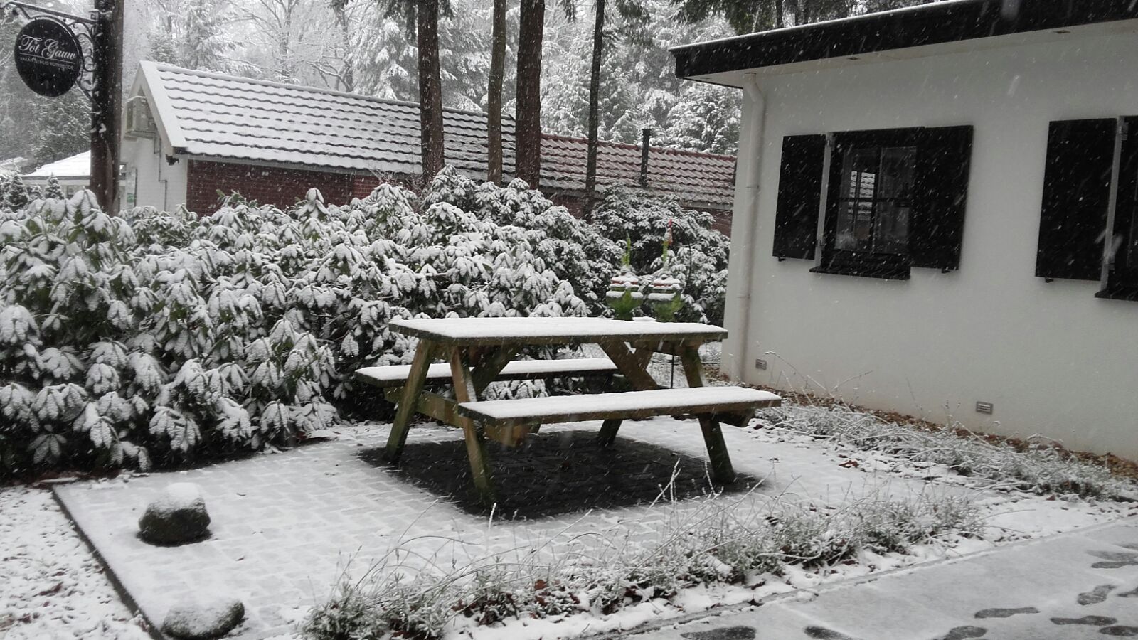 Sneeuwlandschap: Houten picknicktafel en met sneeuw bedekte struiken voor een wit gebouw met zwarte luiken.