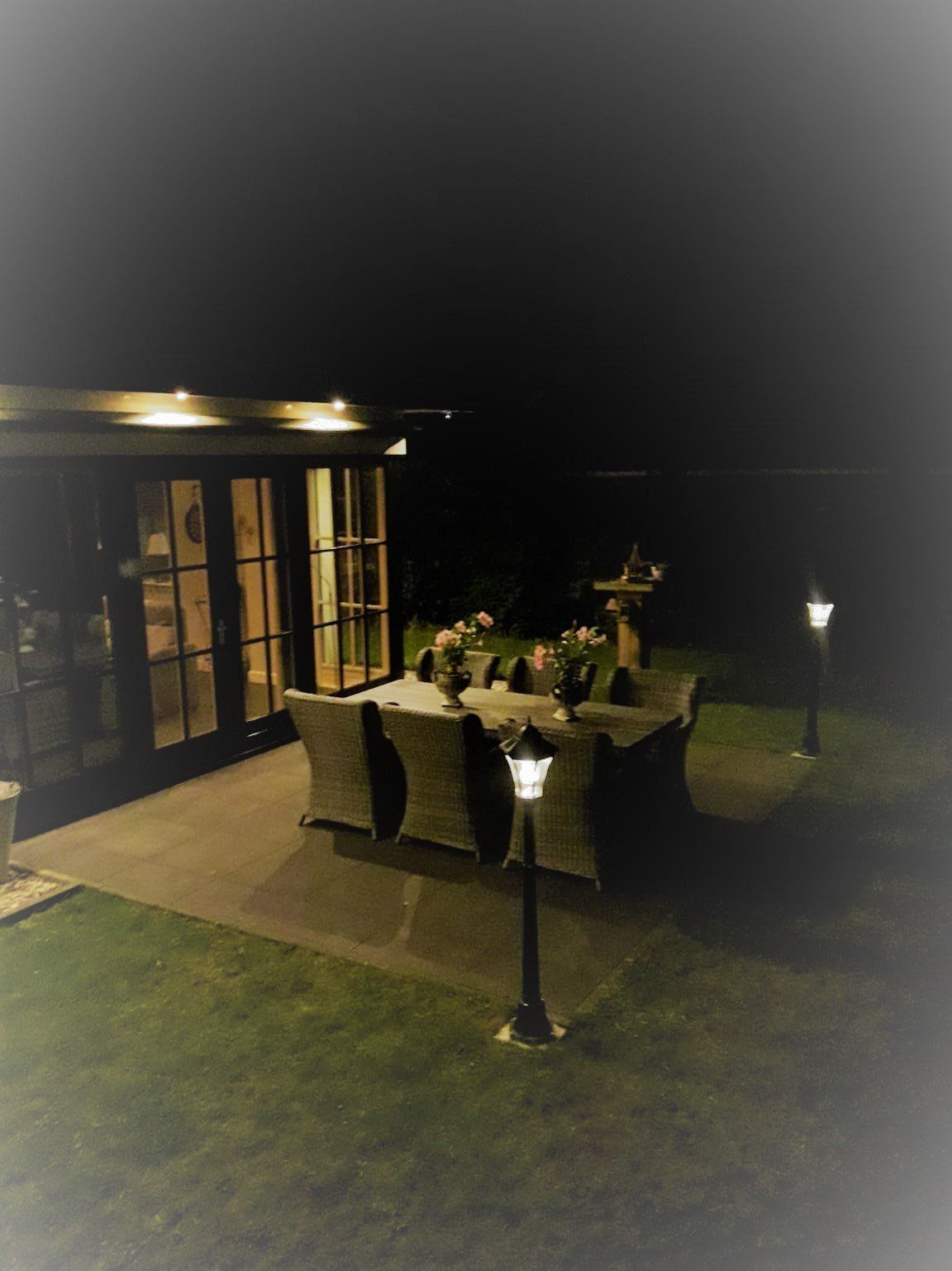 Een terras om 's avonds buiten te dineren, verlicht door een paar lantaarntjes. Een tafel gedekt met bloemen, naast een gebouw met glazen deuren.