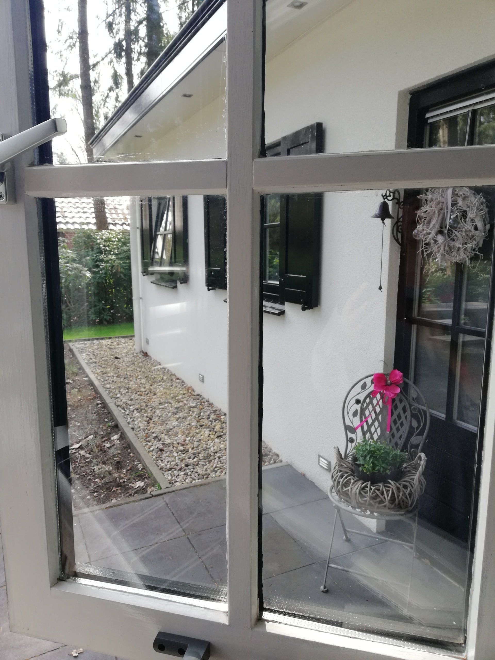 Uitzicht door een raam met witte kozijnen op een wit gebouw met zwarte luiken, een pad en potplanten.