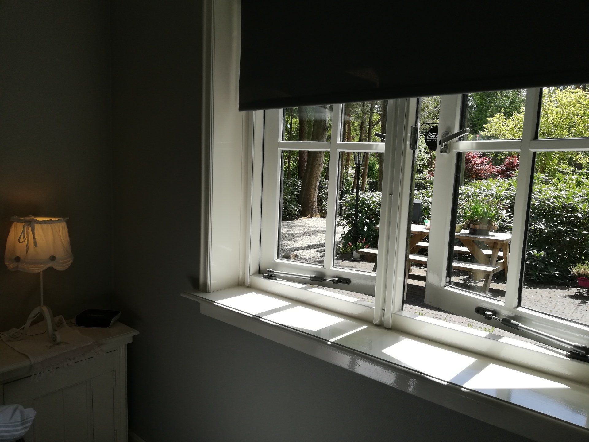 Raam met wit kozijn en een gedeeltelijk opgetrokken zwart rolgordijn. Fel zonlicht valt op de vensterbank en biedt uitzicht op de tuin. Naast het raam staat een lantaarn.