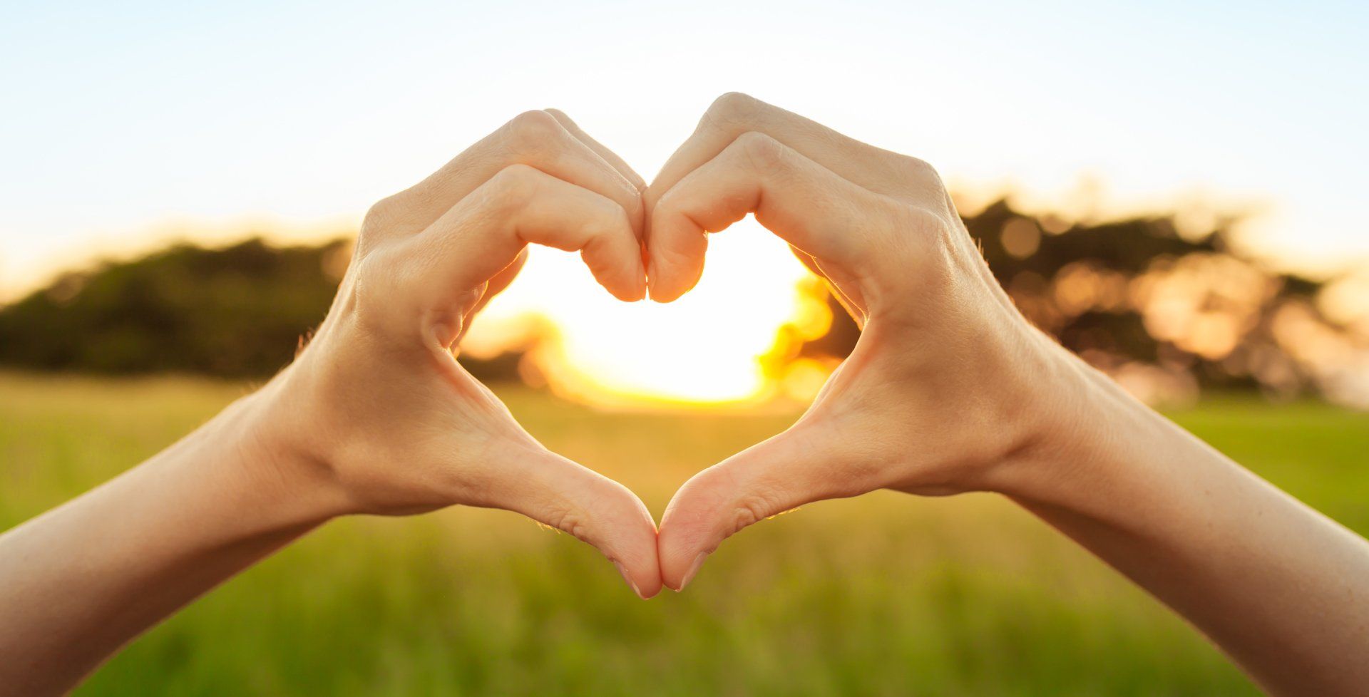 Stock picture of two hands forming a heart shape that frames a blurred sun peeking between the trees at the edge of a field