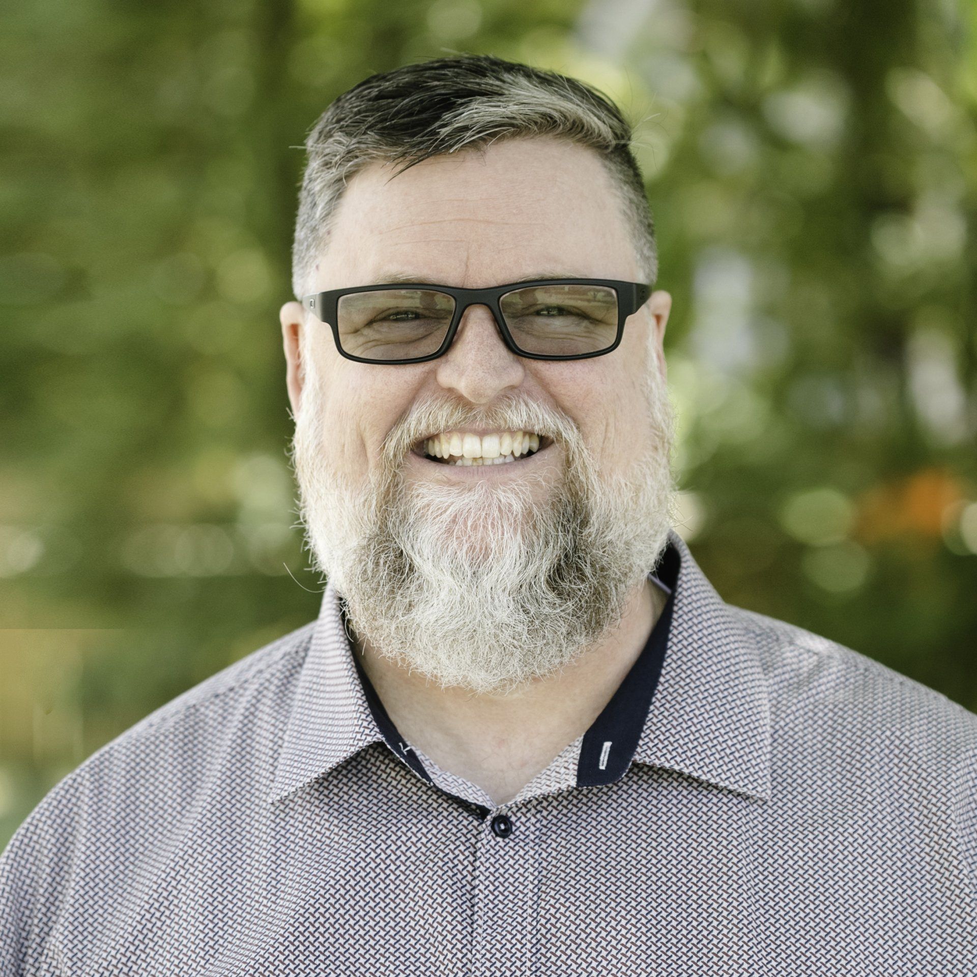Portrait of a bearded man in a gray shirt and black glasses, in front a blurred green background.