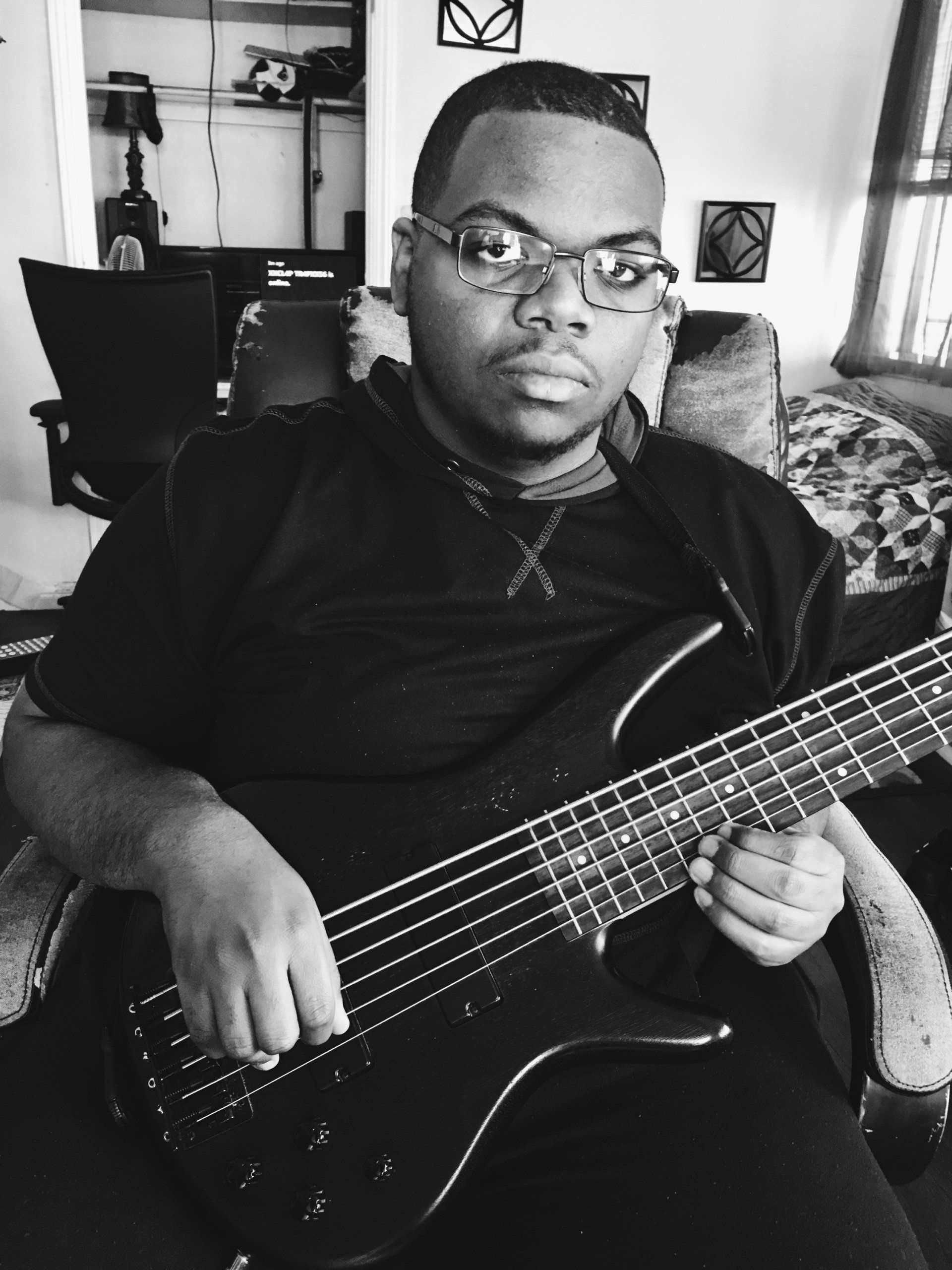 Black and white photo of a man with glasses playing a 6-string bass guitar. He is inside.