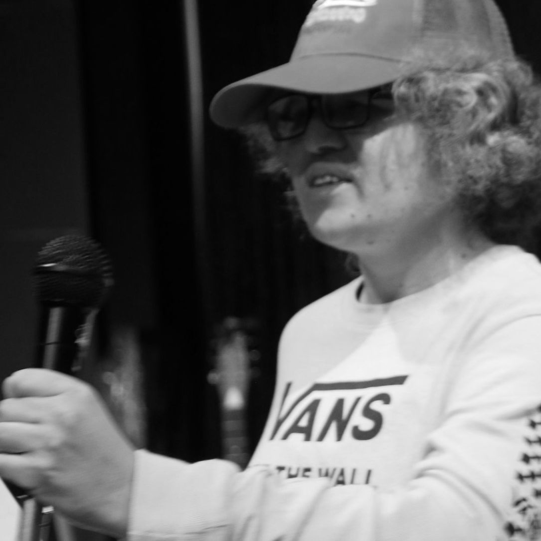 Person with curly hair, wearing a baseball cap and Vans sweatshirt, holding a microphone, speaking.
