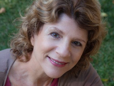 Woman with curly brown hair smiles at camera, wearing pink shirt and tan sweater in a green yard.