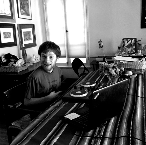 Man smiling at a table with turntables in a room with a window and art on the wall.
