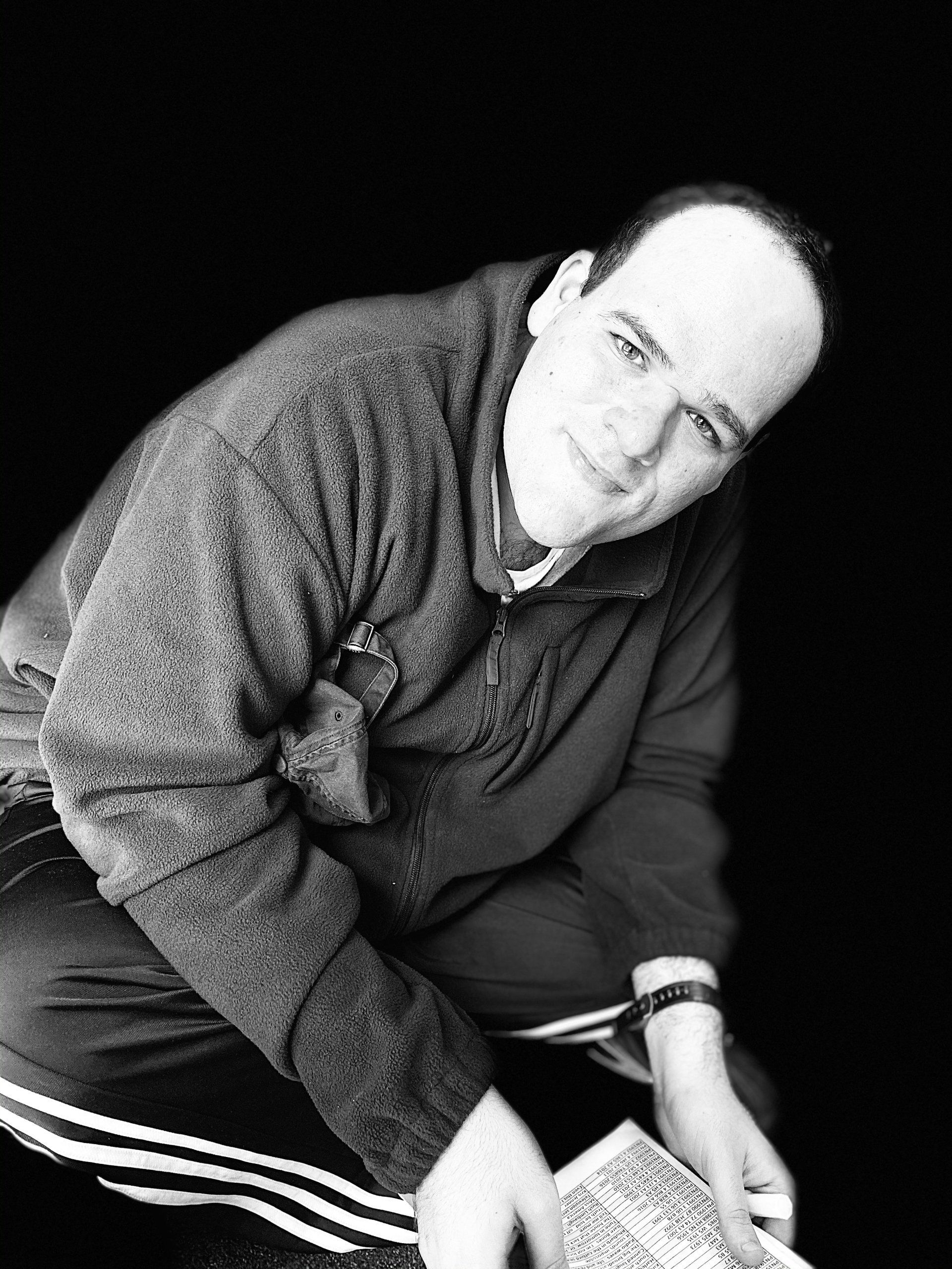 Man in a dark sweater, squatting, holding a paper, looking at the camera, black and white.